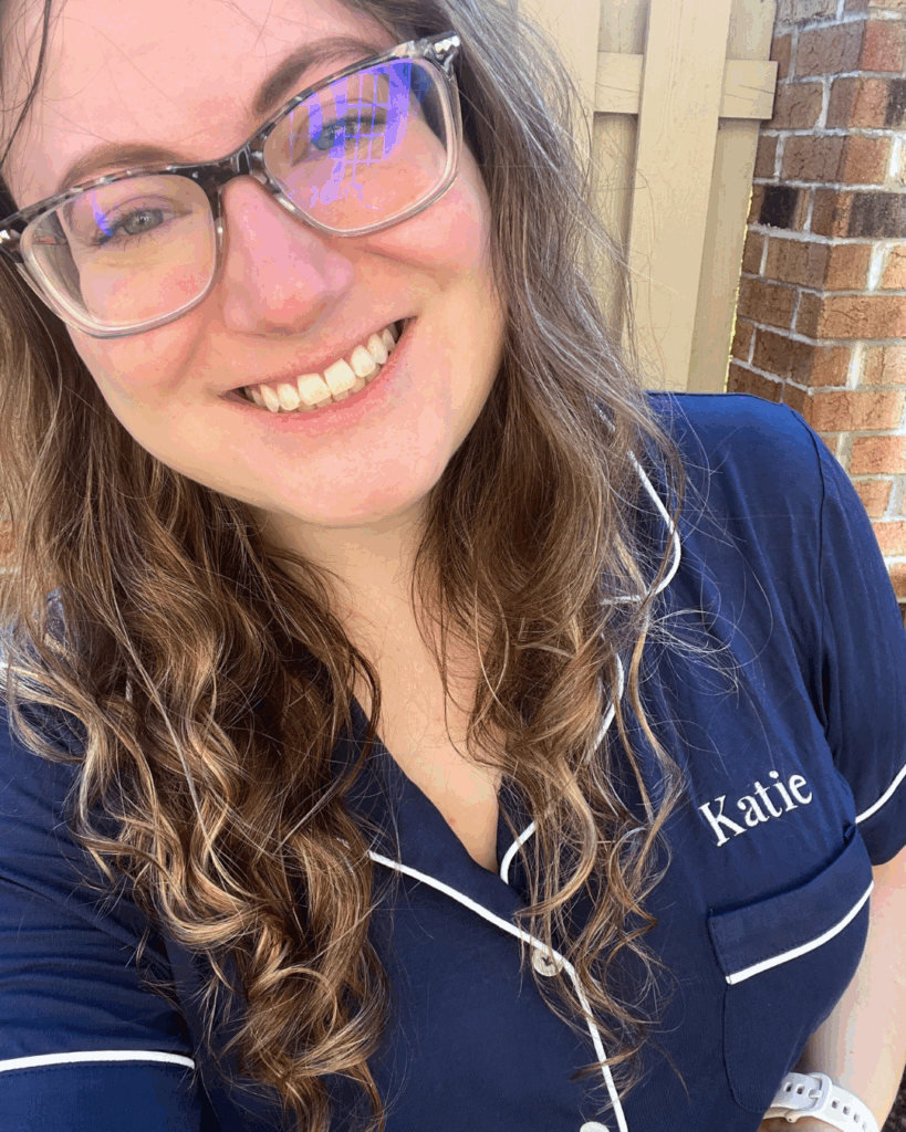 Woman smiling in a navy Birdy Grey pajama set with white piping and personalized name ‘Katie’ embroidered on the pocket.