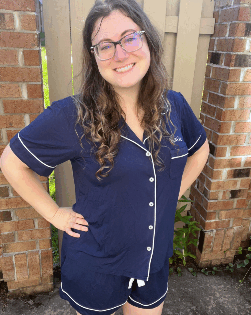 Woman smiling in a navy Birdy Grey Jonny pajama set with white piping and personalized name ‘Katie’ embroidered on the pocket.