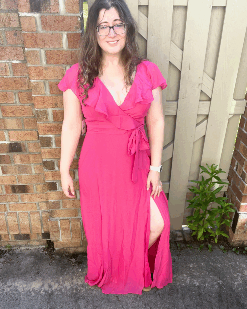 Woman standing in a full-length hot pink, Barbie pink, Birdy Grey Jackson chiffon bridesmaid dress with tie waist and slit, smiling in front of a brick wall and wooden fence.