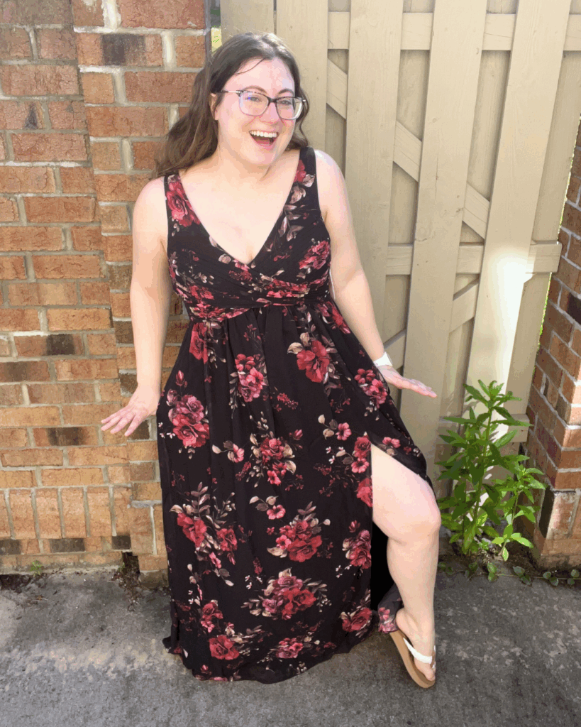 Woman laughing and posing outdoors in a black Birdy Grey Laurie Empire chiffon bridesmaid dress with red and pink floral print and thigh-high slit.