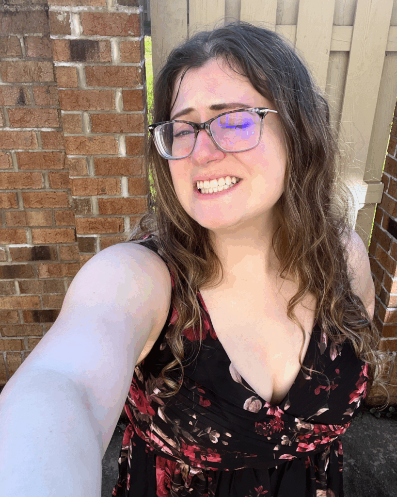 Woman squinting in the sunlight while taking a selfie in a black Birdy Grey Laurie Empire chiffon bridesmaid dress with red floral pattern.