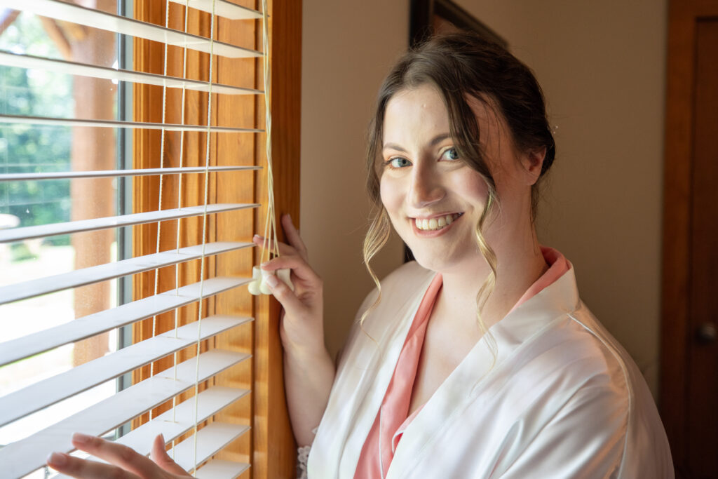 Bride smiling while peeking out the window, wearing white Birdy Grey robe over pink Birdy Grey pj set.