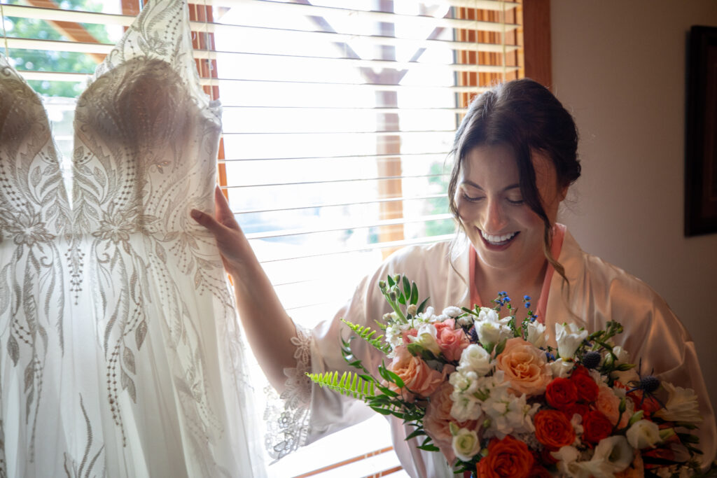 Bride smiling while holding a bouquet of peach, coral, and white flowers, wearing a white Birdy Grey robe over a pink Birdy Grey pajama set, photographed by Cooper’s Photography in North Carolina.