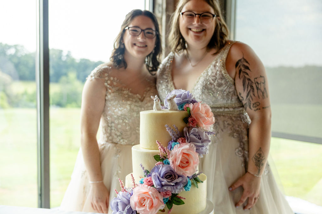 Two Brides in front of their wedding cake, which features non-traditional cake topper (bunnies) and purple, blue, and pink wedding floral details on the cake.